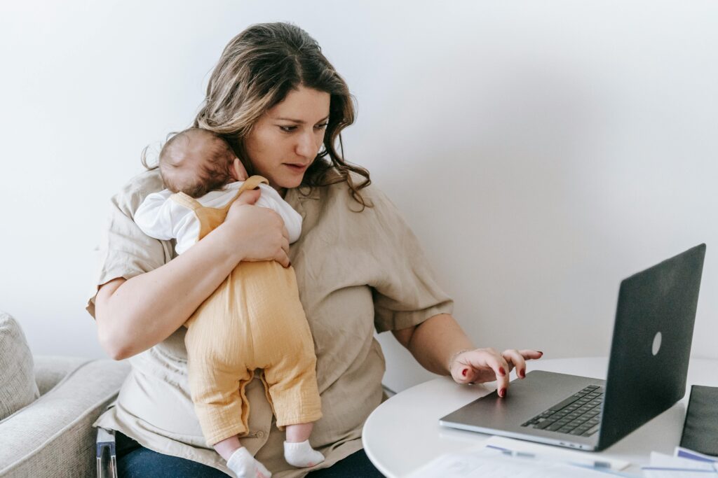 Mother holding baby while working from home on laptop.